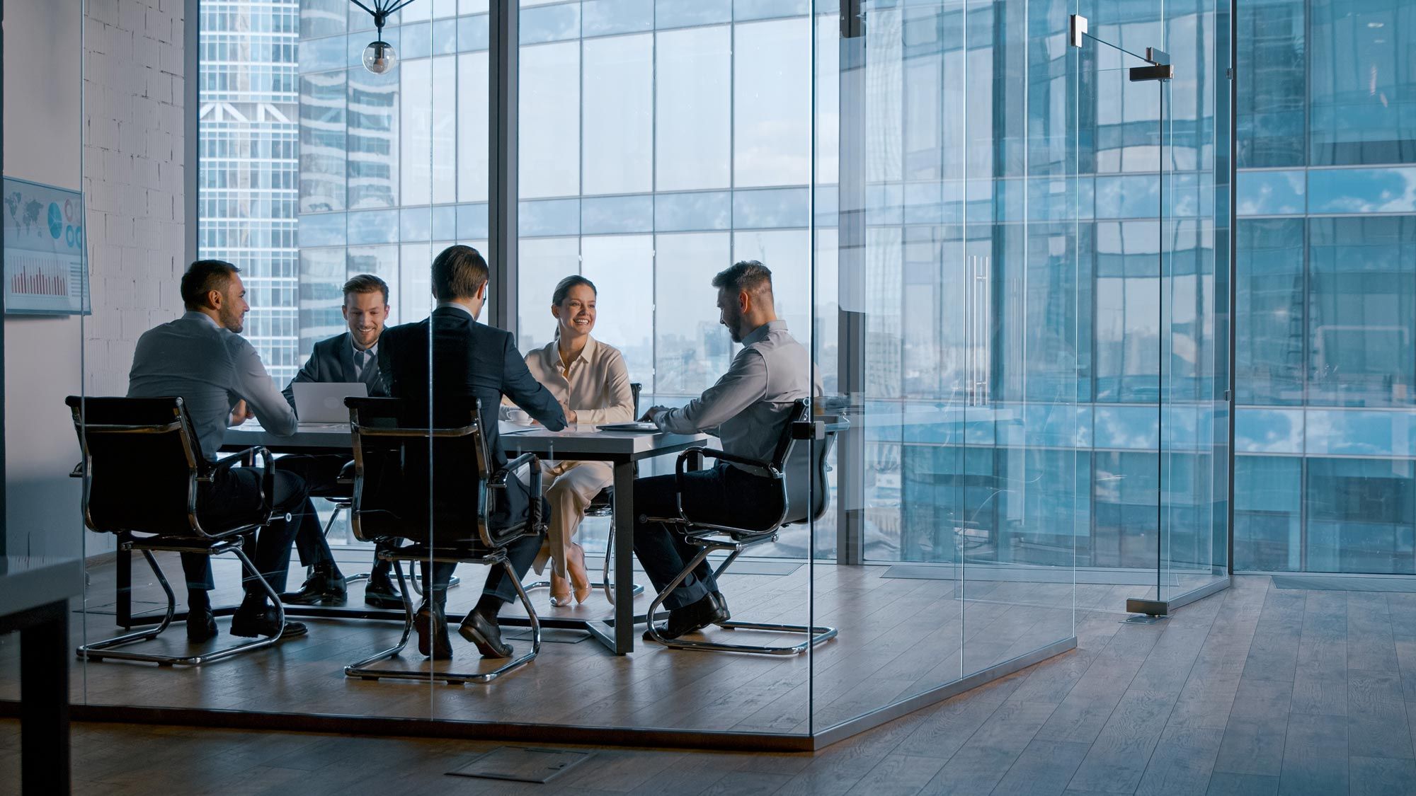 A team meeting around a table in a glass-walled room overlooking office towers